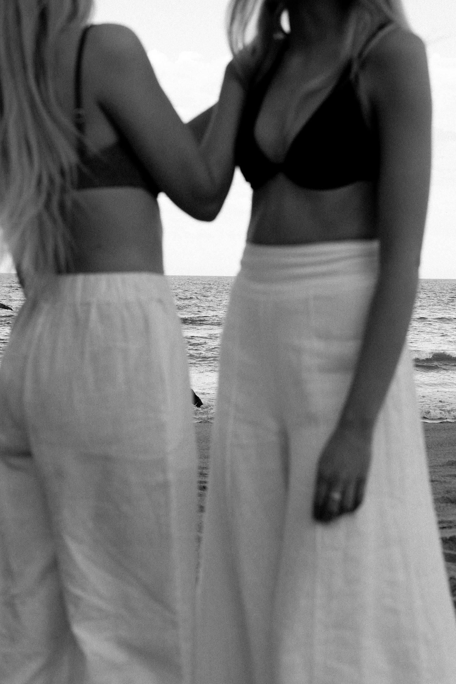 Two women standing close together on a beach, facing away from the camera.